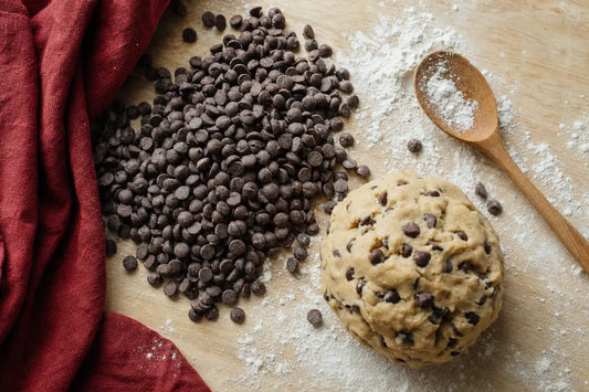 A mound of dark chocolate chips and a ball of cookie dough on a floured wooden board with a wooden spoon