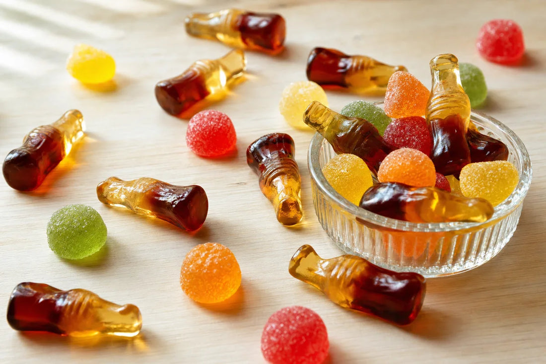 Glossy cola bottle and round fruit jelly sweets scattered across a maple table beside a small glass dish