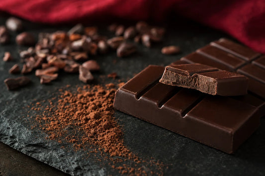 A snapped bar of dark chocolate on slate with cocoa powder and cocoa nibs beside it