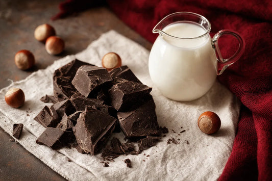 Chunks of dark chocolate on a linen cloth beside a small jug of milk and whole hazelnuts