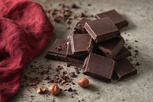 Squares of dark chocolate broken from a bar on a stone surface with cocoa nibs and hazelnuts