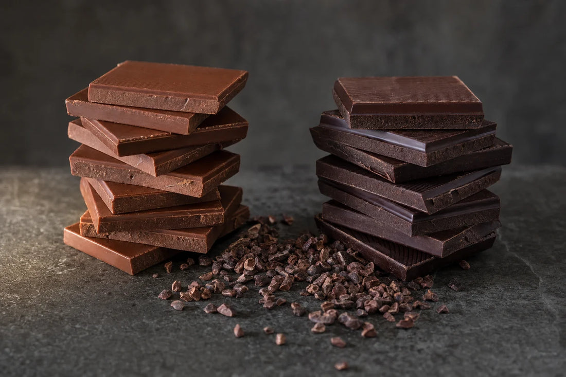Two stacks of chocolate squares side by side on a dark concrete tabletop with cocoa nibs between them