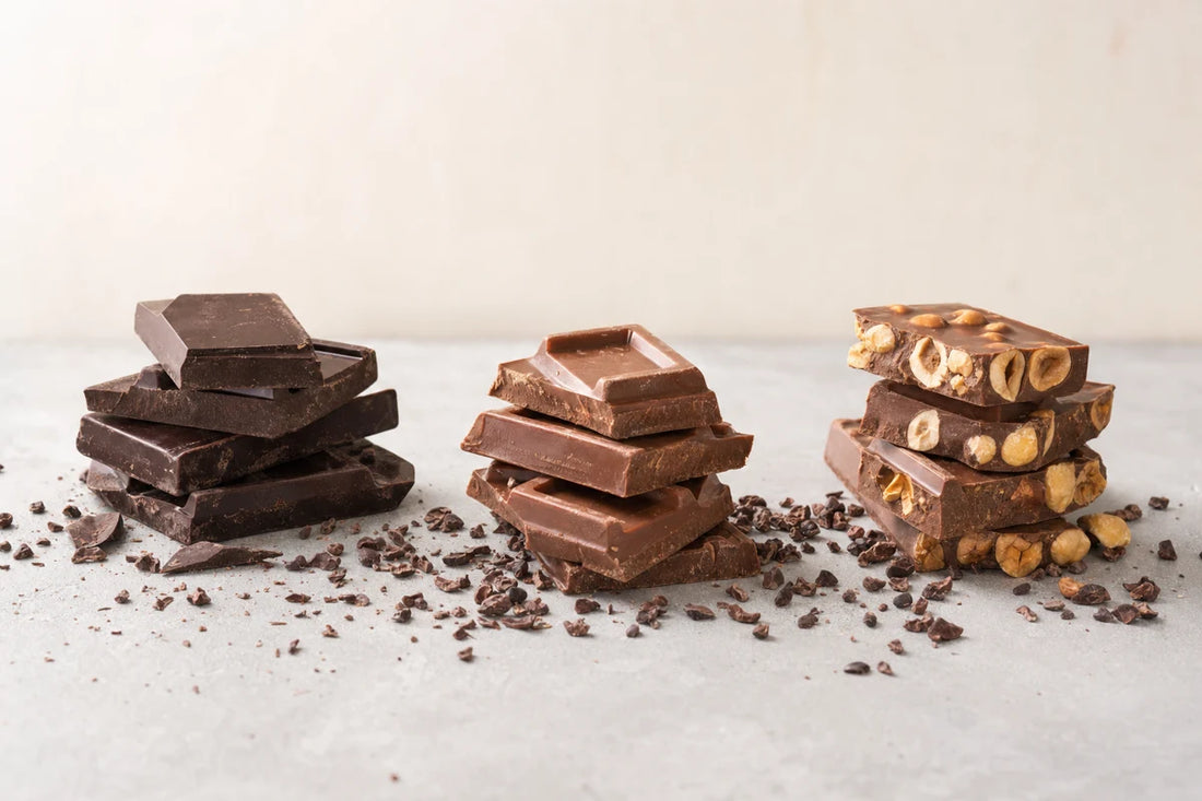 Three groups of dark, milk and hazelnut chocolate pieces in a row on a concrete tabletop with cocoa nibs