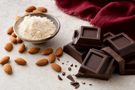 Squares of dark chocolate on pale stone beside a bowl of almond flour and a scatter of whole almonds