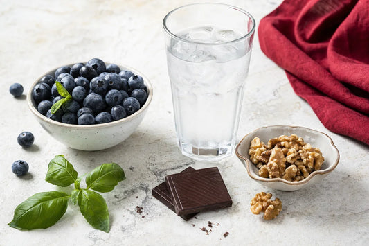 A glass of iced water, a bowl of blueberries, walnuts and two squares of dark chocolate on a pale stone counter