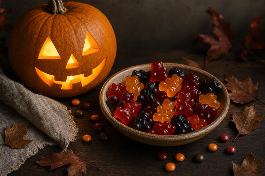 A bowl of glossy red, orange and dark purple gummy bears beside a lit carved pumpkin on a dark wooden table