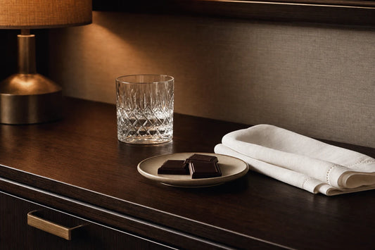 A cut glass tumbler, a folded linen napkin and a plate of dark chocolate squares on a hotel console table