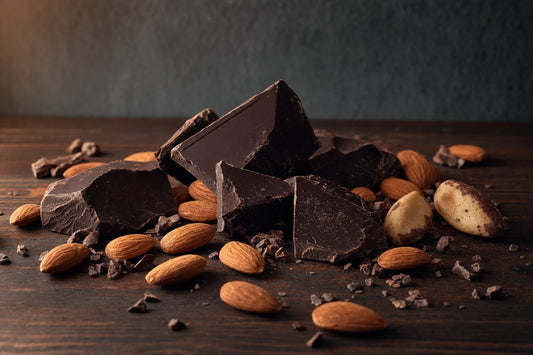 Broken pieces of very dark chocolate on a walnut table with almonds, brazil nuts and cocoa nibs