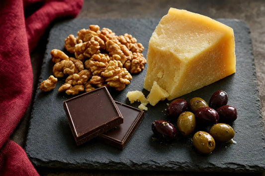 A slate board holding walnuts, a wedge of hard cheese, dark chocolate squares and olives