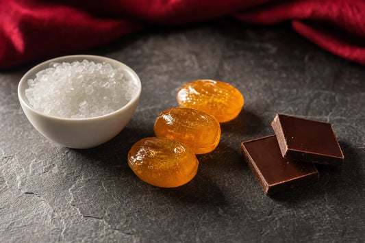A bowl of white sweetener crystals on slate beside three amber boiled sweets and two squares of dark chocolate
