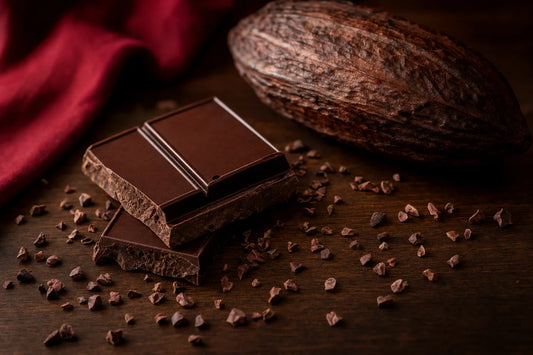 A snapped bar of dark chocolate on a walnut table with cocoa nibs scattered around it