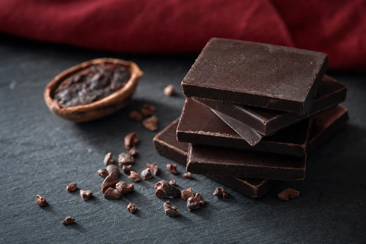 A stack of dark chocolate squares on slate with cocoa nibs scattered around them