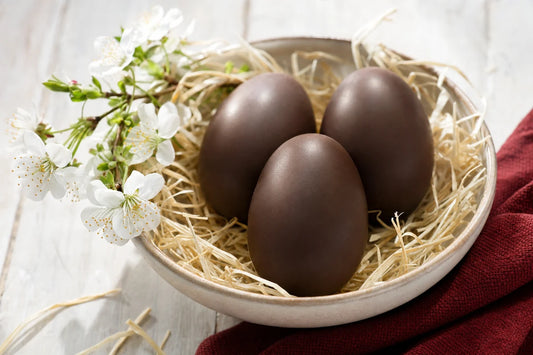 Three plain dark chocolate eggs nestled in straw in a ceramic bowl beside a sprig of spring blossom