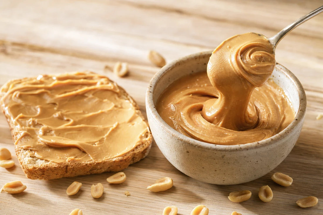 A bowl of smooth peanut butter with a spoon lifting a ribbon of it beside toast and shelled peanuts