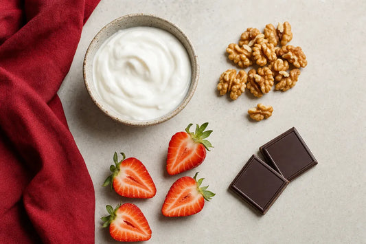 A bowl of plain yoghurt, walnuts, halved strawberries and two squares of dark chocolate on a pale stone surface