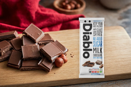 Diablo No Added Sugar Milk Chocolate 85 g bar standing on an oak board beside broken milk chocolate squares and two hazelnuts