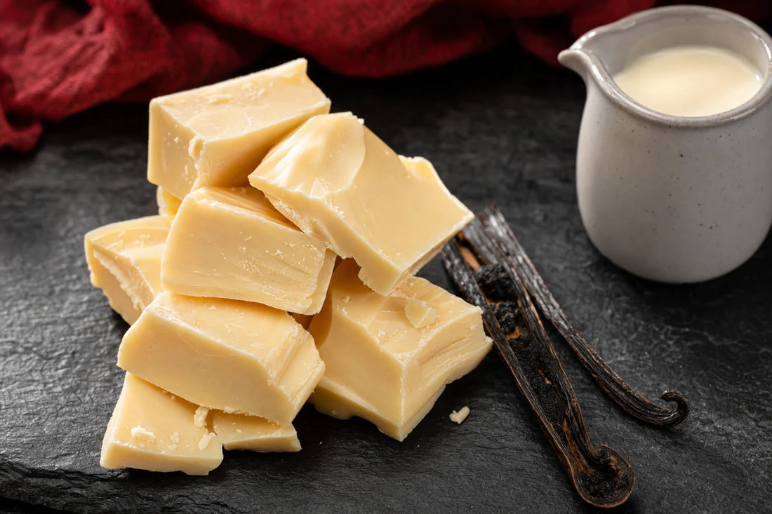 Chunks of white chocolate stacked on dark slate beside a split vanilla pod and a small jug of cream