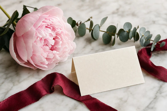 A pink peony and eucalyptus on a marble surface beside a blank cream card and deep red silk ribbon