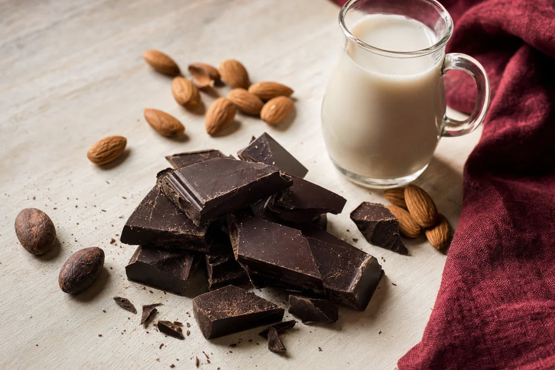 Broken pieces of dark chocolate on a pale wooden board beside almonds, cocoa beans and a small jug of plant milk