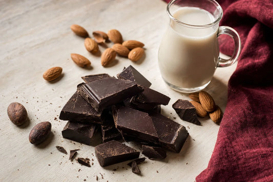 Broken pieces of dark chocolate on a pale wooden board beside almonds, cocoa beans and a small jug of plant milk