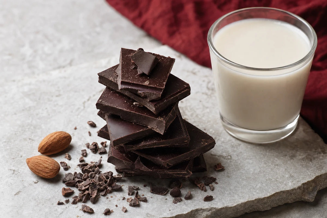 A stack of very dark chocolate pieces on a stone slab beside a glass of oat milk, cocoa nibs and almonds