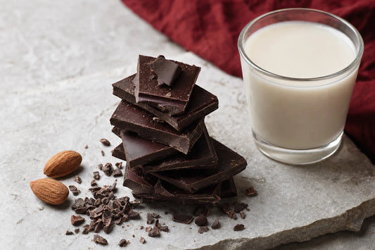 A stack of very dark chocolate pieces on a stone slab beside a glass of oat milk, cocoa nibs and almonds