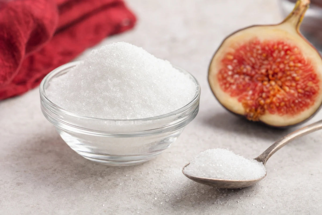 A glass bowl of fine white sweetener crystals on pale concrete beside a spoonful and a halved fresh fig