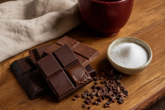 Broken squares of dark and milk chocolate beside a small bowl of white crystalline maltitol and scattered cocoa nibs on an oak board